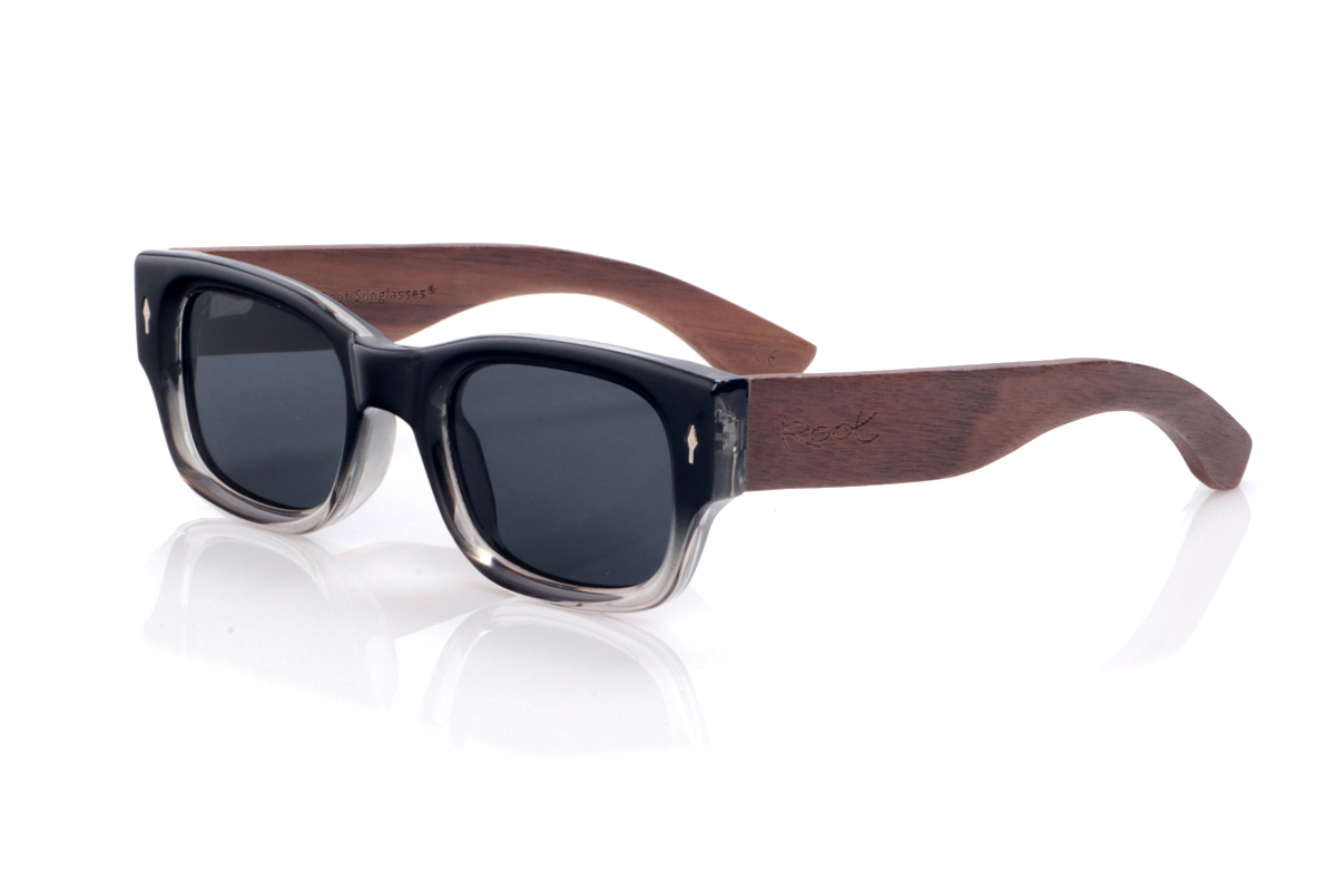 Gafas de Madera Natural de Walnut modelo NEEL. Las gafas de sol <b>NEEL</b> combinan un frontal grueso en negro con degradado a transparente en la parte inferior y pequeños apliques metálicos en forma de flecha en las esquinas, que refuerzan su aire retro y contundente. La forma rectangular suavemente redondeada ofrece un ajuste cómodo en rostros medianos, mientras que las varillas anchas de madera de nogal muestran unas vetas cálidas y marcadas que aportan carácter artesanal y un contraste elegante con el frontal. Ligeras pese a su presencia, se sujetan con firmeza y montan lentes polarizadas oscuras que reducen reflejos y protegen eficazmente frente a los rayos UV. Medidas: 144x44 mm. Calibre 52. | Root Sunglasses® 