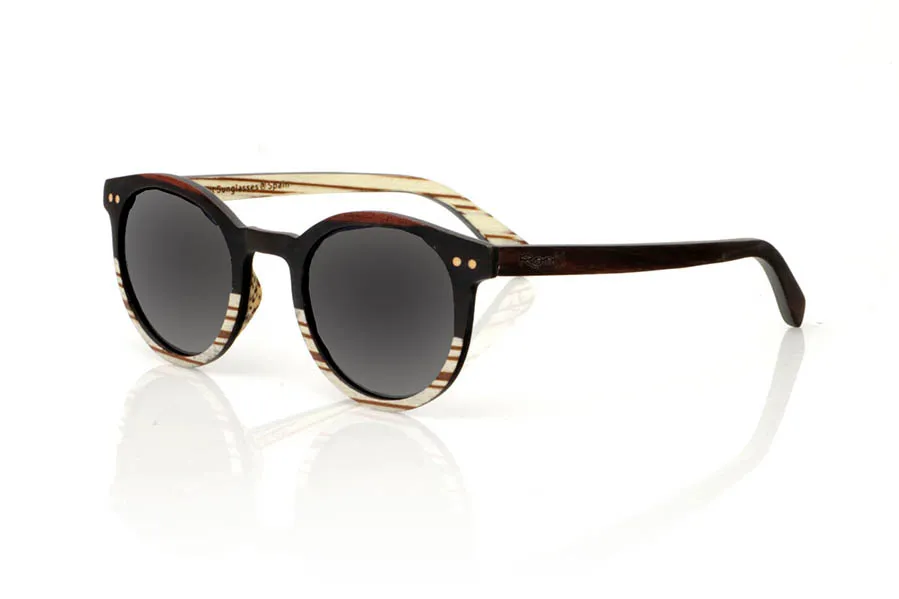Gafas de Madera Natural de Ébano modelo ATHENEA. Gafas de sol de madera ATHENEA, con su diseño compacto y redondeado y una ceja pronunciada similar a los modelos LANA y LINDA, se destacan por su elegancia y originalidad. Fabricadas con una combinación única de madera de ébano en la parte superior del frontal y zebra blanca en la inferior, estas gafas ofrecen un contraste visual impresionante que capta la atención. Además, el frontal se enriquece con dos incrustaciones redondas de madera de haya, añadiendo un detalle distintivo y un toque de sofisticación. Con una medida de 135x47mm y un calibre de 45, las ATHENEA son ideales para quienes buscan gafas más pequeñas sin sacrificar el estilo ni la presencia.  | Root Sunglasses® 