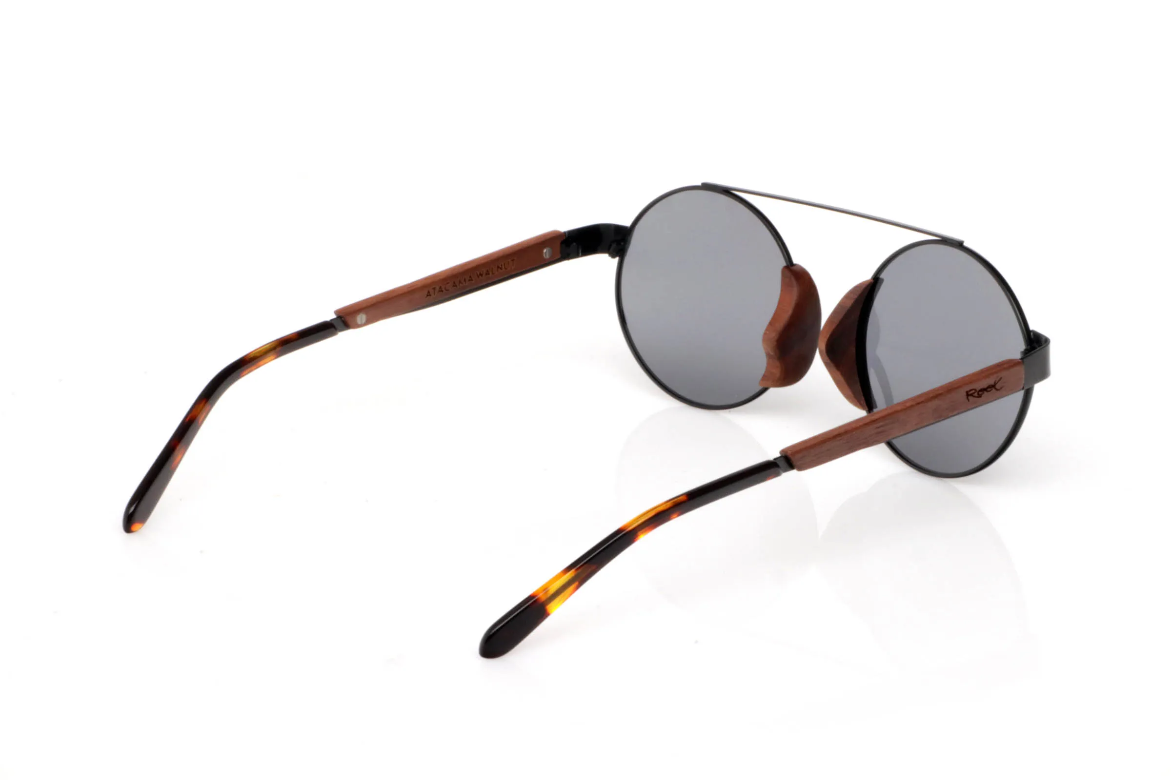 Wood eyewear of Walnut ATACAMA SK. ATACAMA SK wooden sunglasses combine a fine metal structure with walnut details that bring warmth and character. The contrast between black metal and natural wood creates a unique balance: visually light, yet full of personality.

The round shape with a top bridge recalls classic aviator-inspired forms, reinterpreted in a minimal and modern way. The walnut inserts on the front, integrated around the lenses, add a distinctive and original touch without being excessive.

They are especially comfortable thanks to their lightweight construction and flexible feel, making them ideal for long wear. The temples combine wood with tortoiseshell-style tips, adding an extra natural touch.

A subtle yet unique model, perfect for those looking for something different while keeping it simple.

Full UV protection.

Size: 128x49
Lens width: 47 for Wholesale & Retail | Root Sunglasses® 